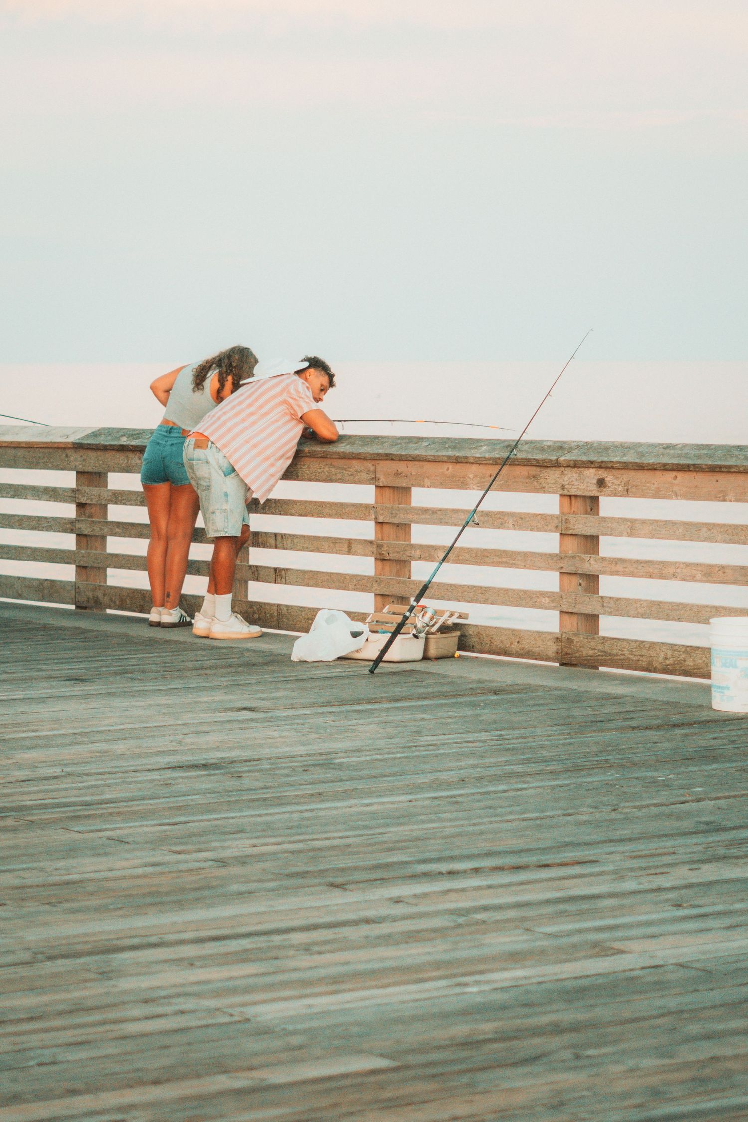 Two people leaning over a wooden pier railing while fishing at sunset near the ocean.