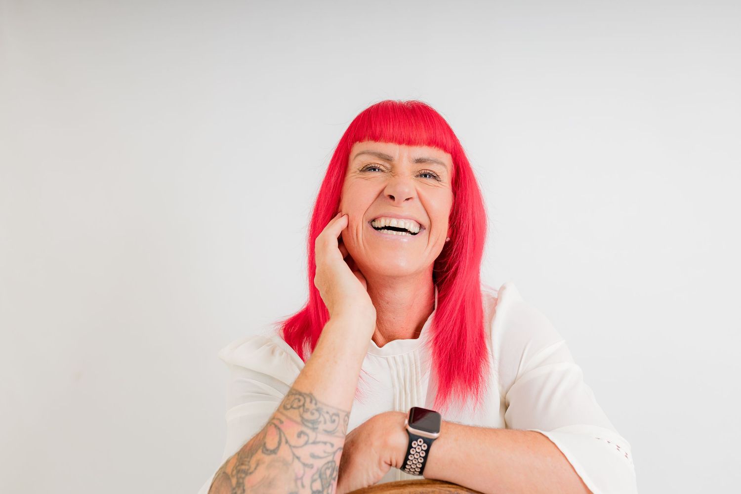 Laughing woman with vibrant pink hair, tattoos, and a smartwatch wearing a white sweater against a light background.