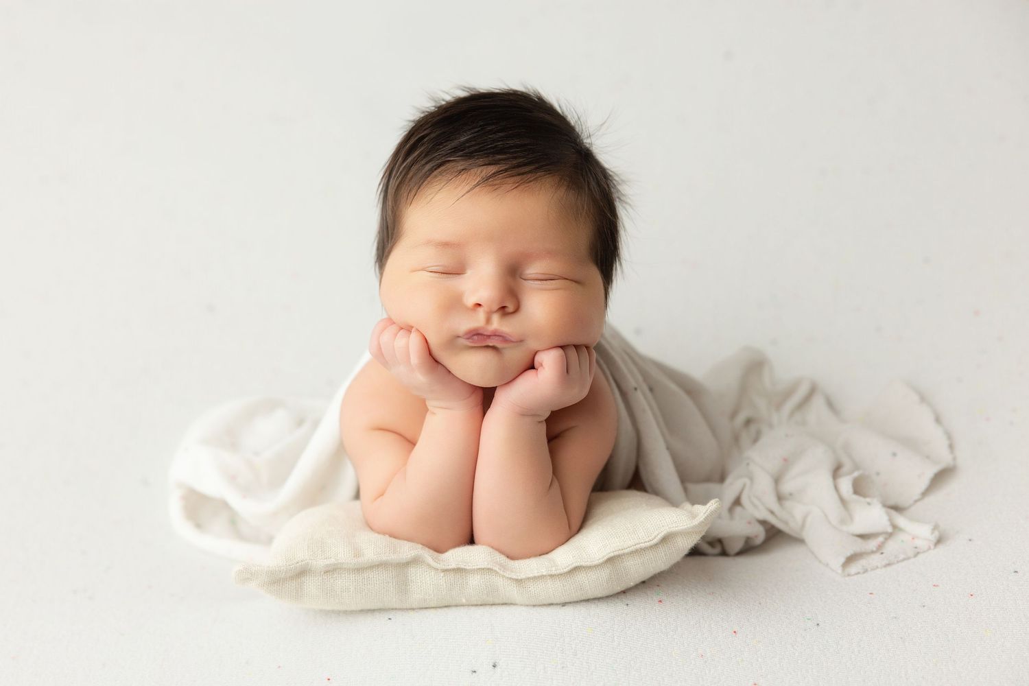 Peaceful newborn baby sleeping with hands under chin on white pillow, wrapped in soft cream fabric.