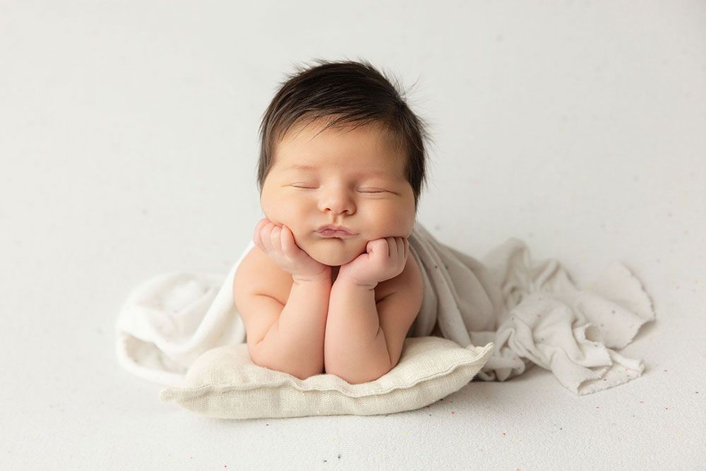 Peaceful newborn baby sleeping with hands under chin on white pillow, wrapped in soft cream fabric.