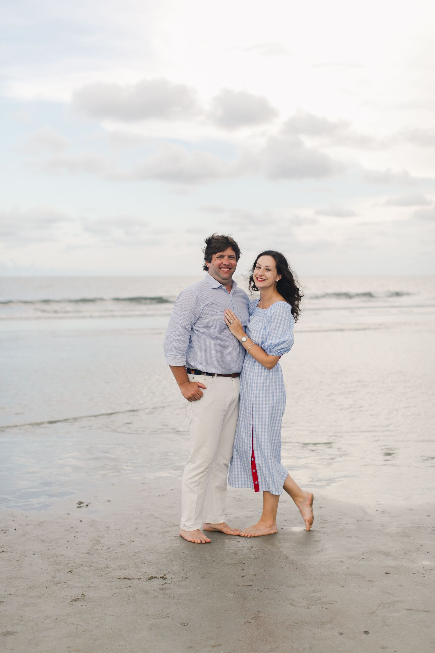 Couple smiling and embracing barefoot on a sandy beach with ocean waves and cloudy sky backdrop at a Beaufort, SC beach shore.