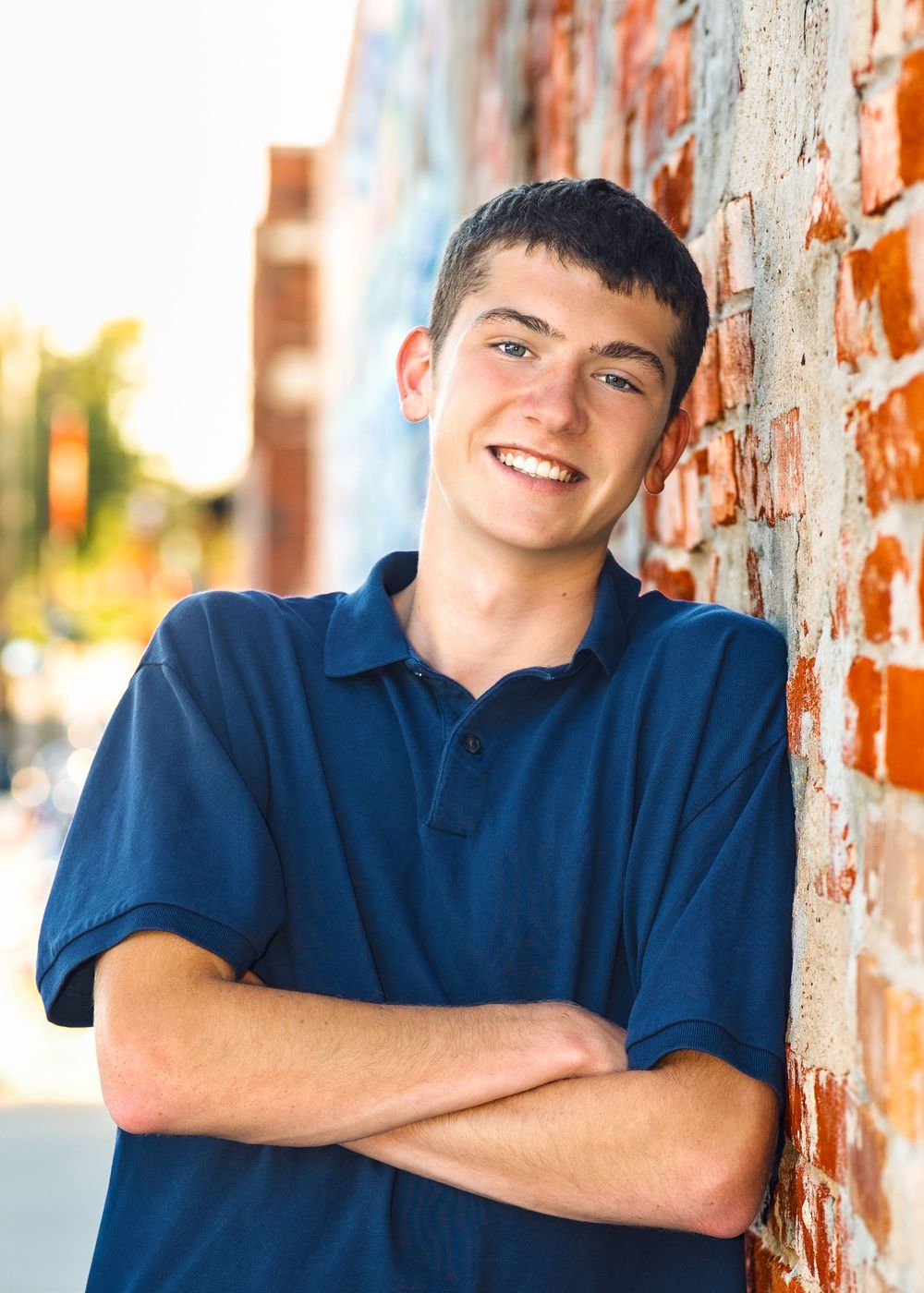 Smiling teenage boy in blue polo shirt leaning against brick wall with arms crossed outdoors.