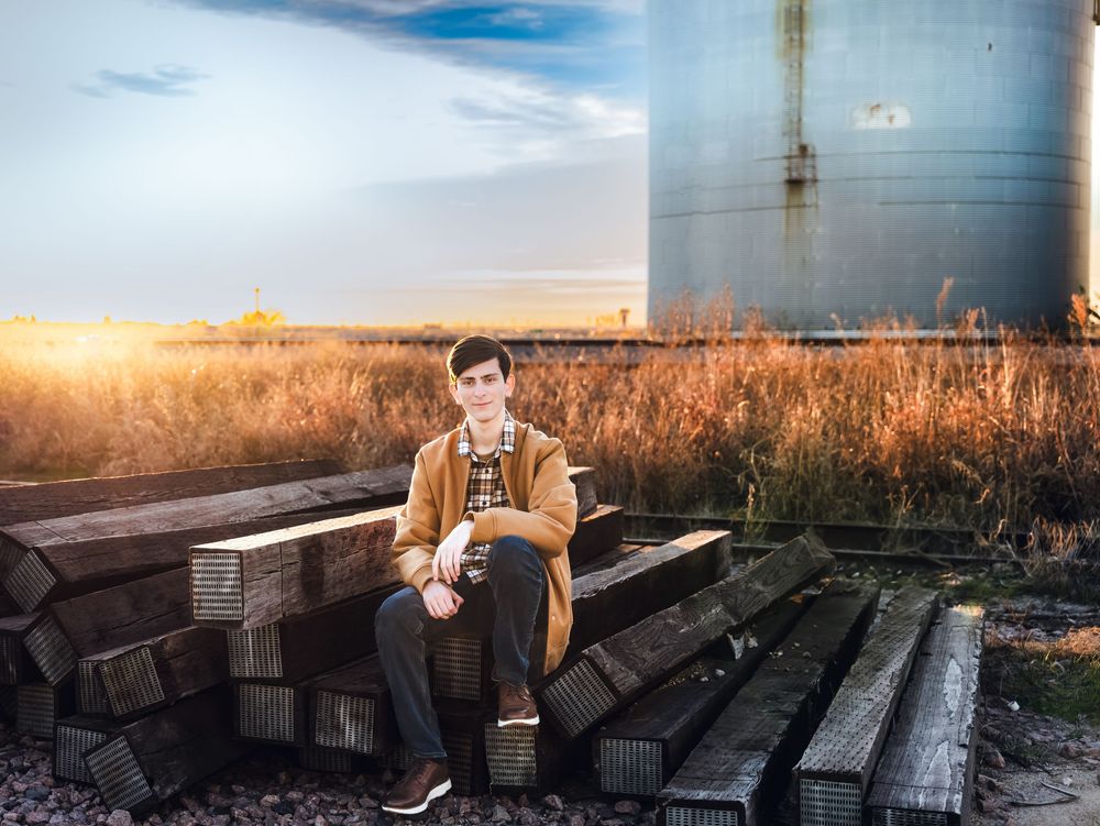 Young man in brown jacket sitting on railroad ties near grain silos at golden hour sunset.