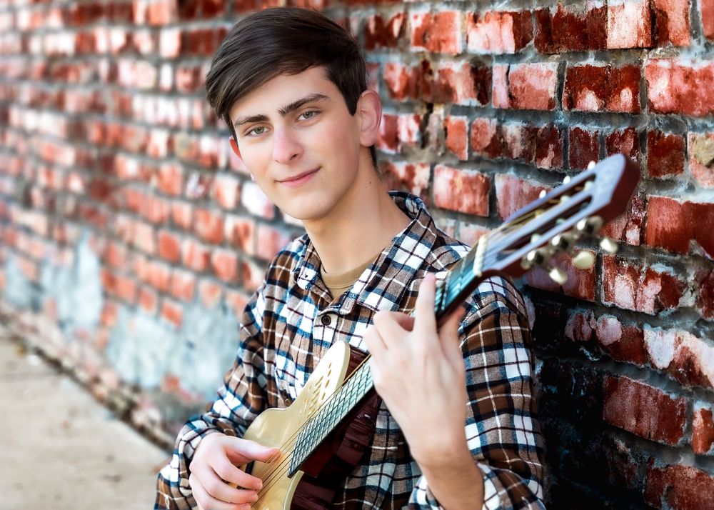 Young man in plaid shirt playing ukulele against a red brick wall outdoors.