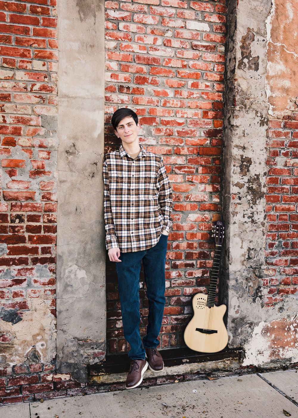 Young man in plaid shirt leaning against rustic brick wall with acoustic guitar, ideal for music or lifestyle photography.