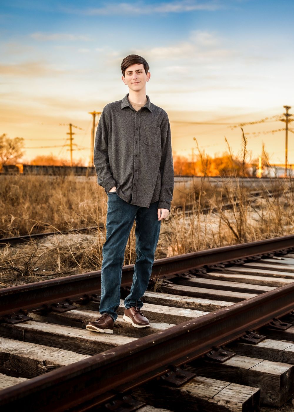 Young man in gray shirt and blue jeans standing confidently on railroad tracks at golden hour sunset.