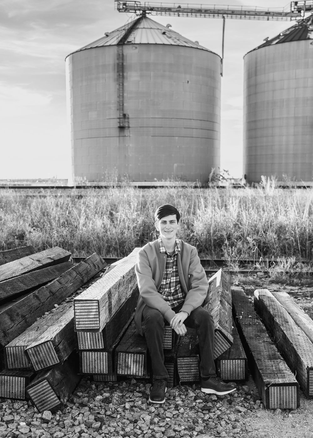 Young man sitting on stacked railroad ties in front of large grain silos, black and white portrait photo.