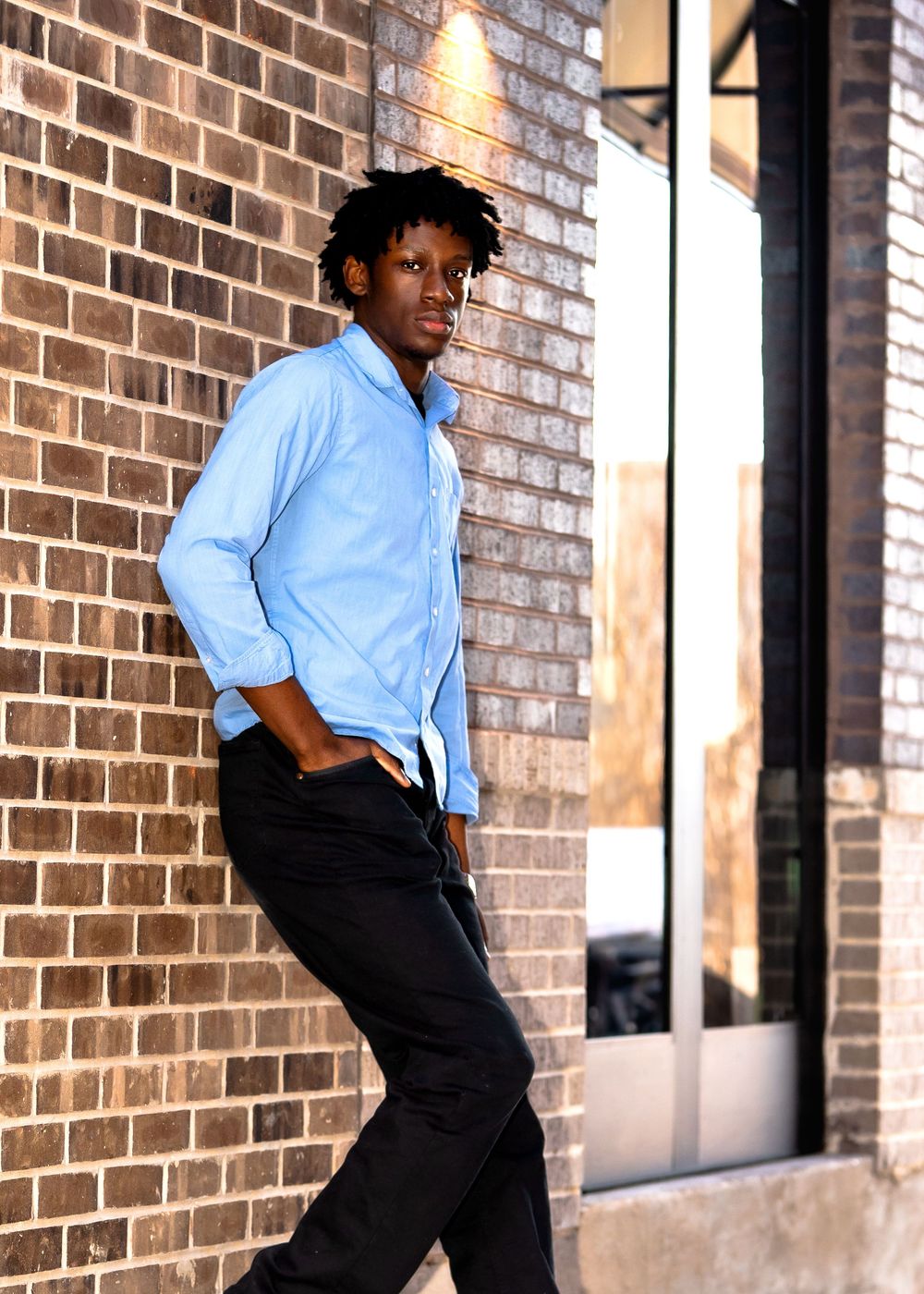 Young man in blue shirt and black pants leaning against a brick wall outdoors.