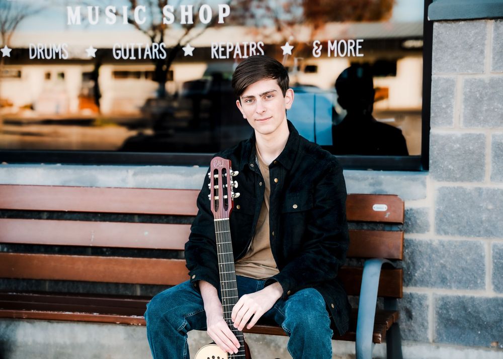 Young man sitting on a bench outside a music shop holding an acoustic guitar.
