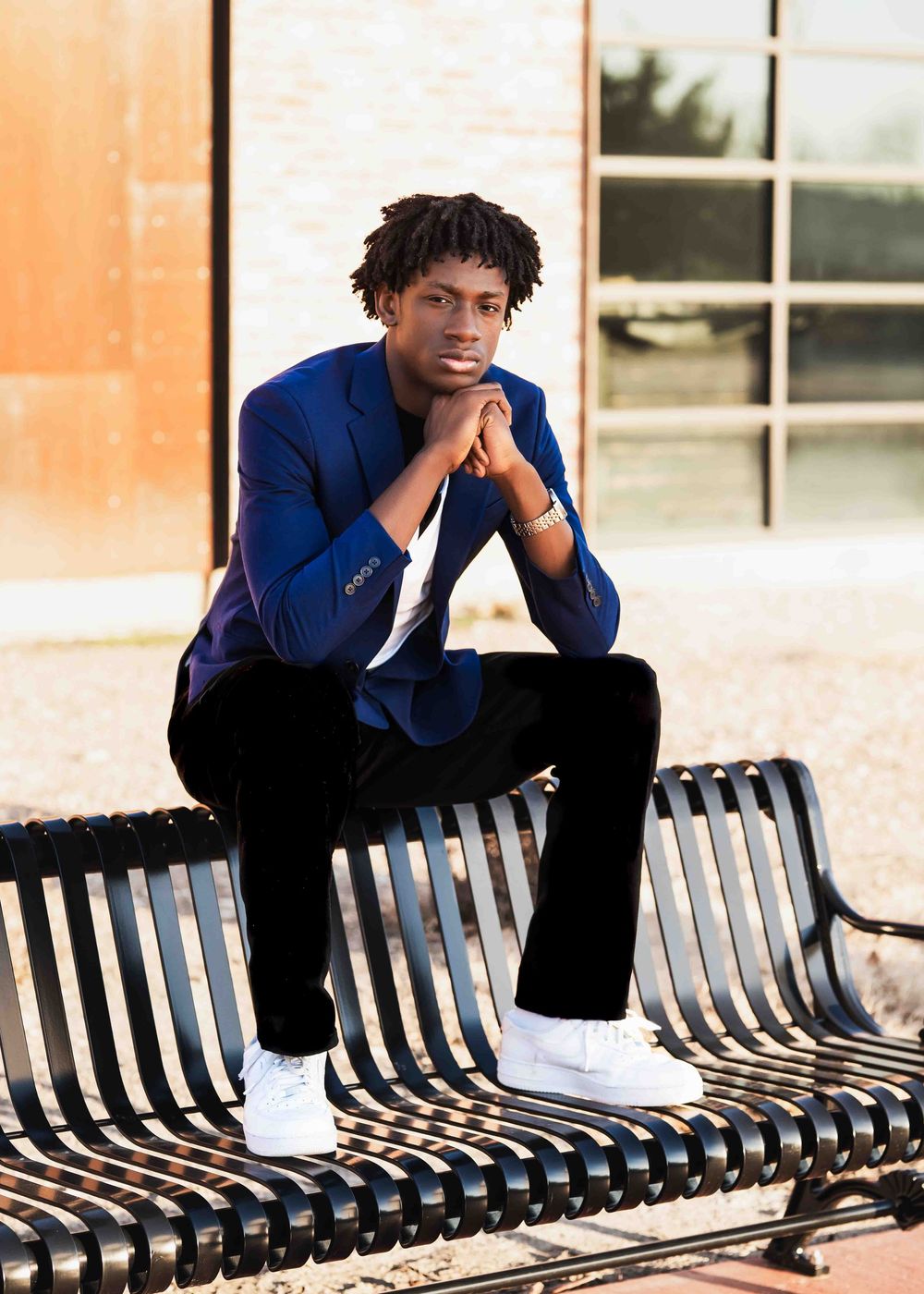 Young man in blue blazer and white sneakers sits on a bench outdoors, posing thoughtfully with hands clasped.