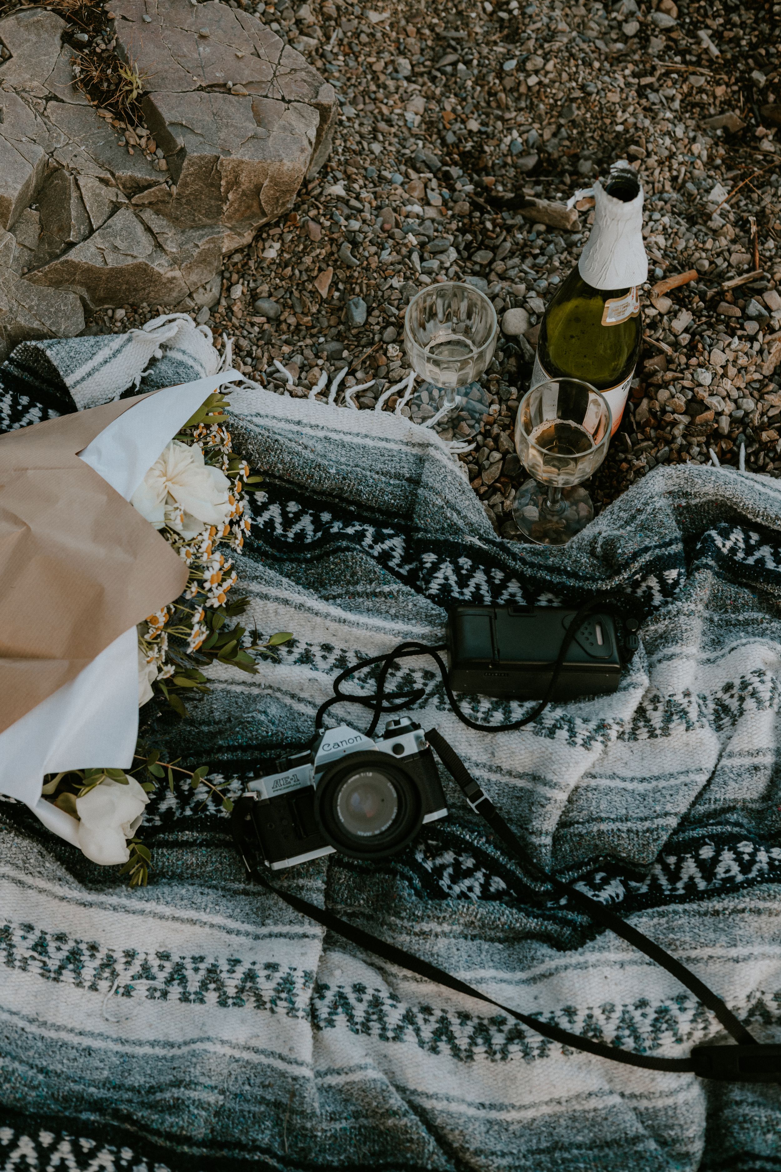 Rustic picnic blanket with wine bottle, glasses, camera, and floral bouquet on rocky ground.
