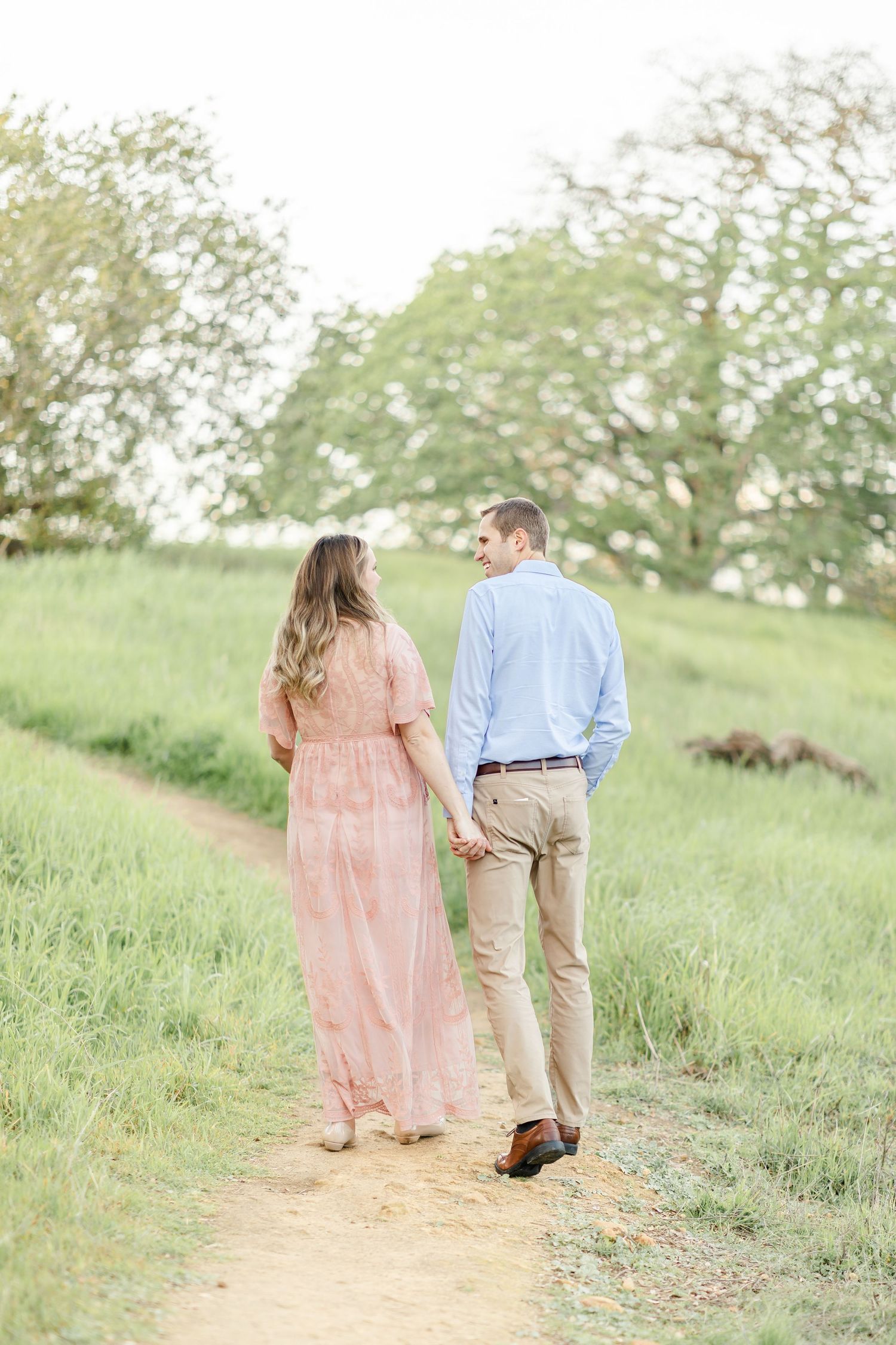 Pregnant couple sharing a kiss outdoors, woman in pink dress cradling baby bump, man in blue shirt, golden hour light.