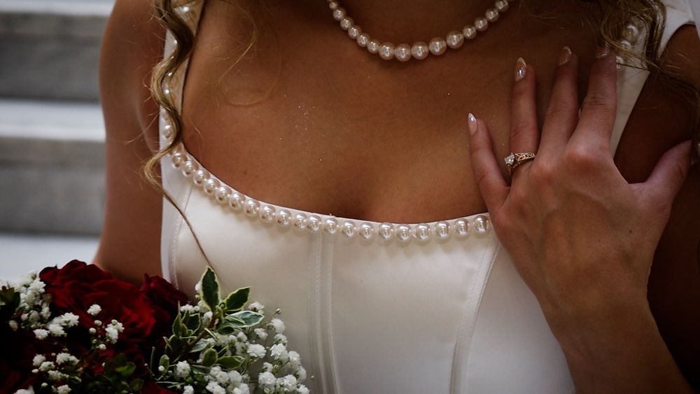Bride in white satin wedding dress with pearl necklace holding red roses bouquet, wedding ring visible on hand.
