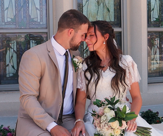 Bride and groom sharing a romantic moment, woman holding bouquet, man in tan suit, seated outdoors near stained glass windows.