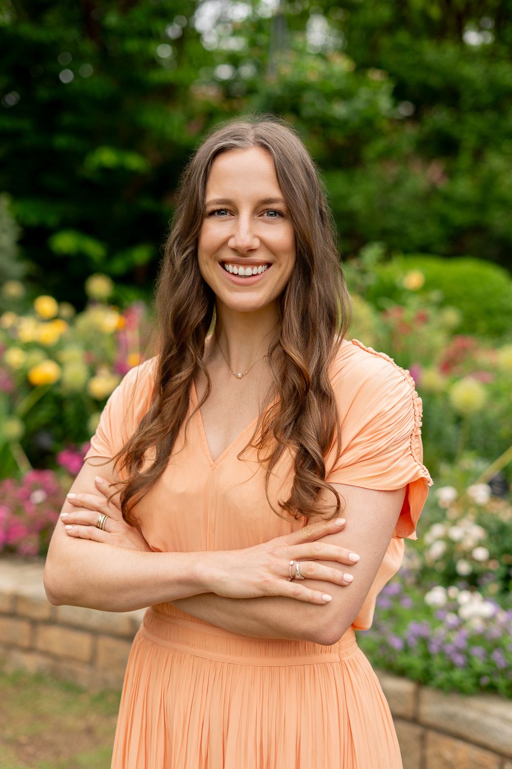 Smiling woman in peach dress with arms crossed standing in colorful garden with flowers.