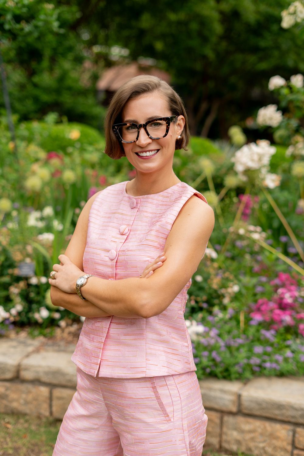 Stylish woman in pink outfit and glasses smiling with arms crossed in blooming garden.