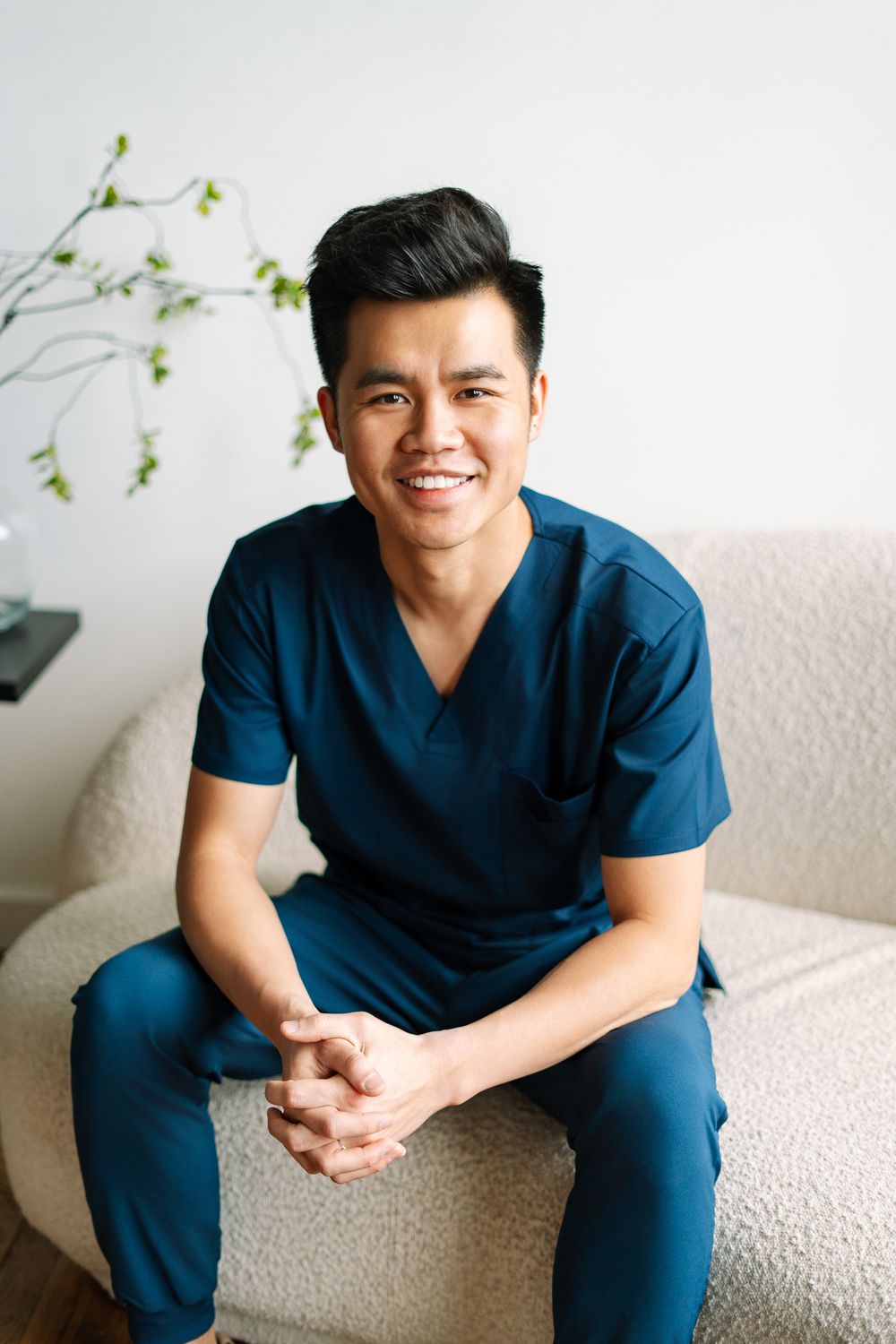 Smiling man in blue scrubs sitting on cream sofa with hands clasped in bright white room.
