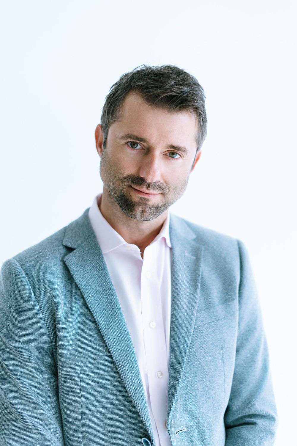 Professional man with beard wearing grey blazer posing against light background.
