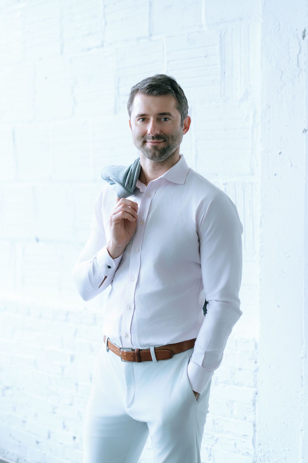 Confident man in white shirt and pants holding jacket over shoulder against white brick wall.