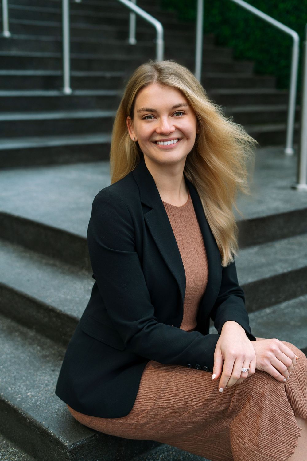 Smiling blonde woman in black blazer sitting on outdoor steps with wind-blown hair.