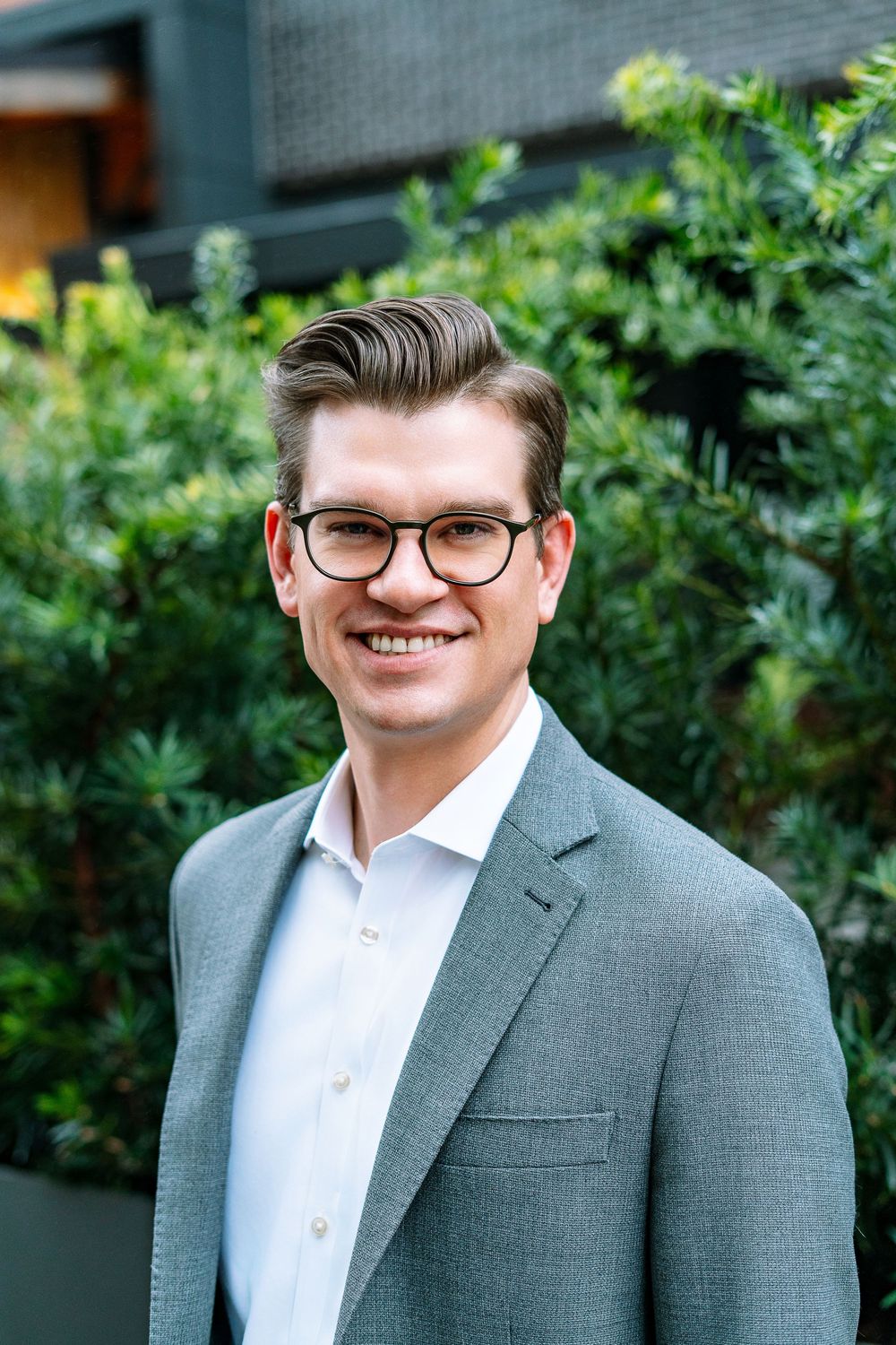 Professional man in gray blazer and glasses smiling outdoors with green foliage background.