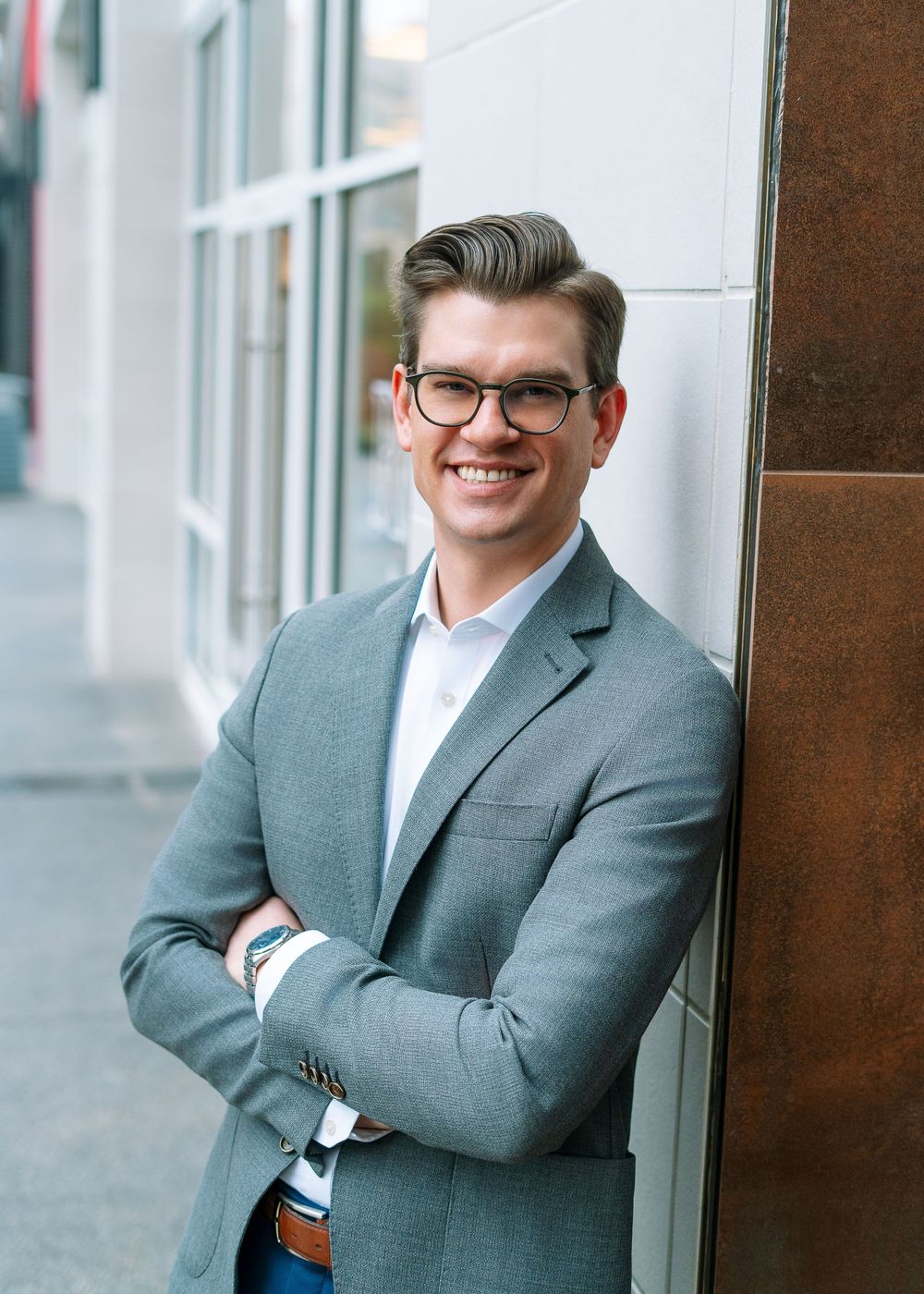 Smiling man in gray blazer and glasses leaning against wall with arms crossed on city street.
