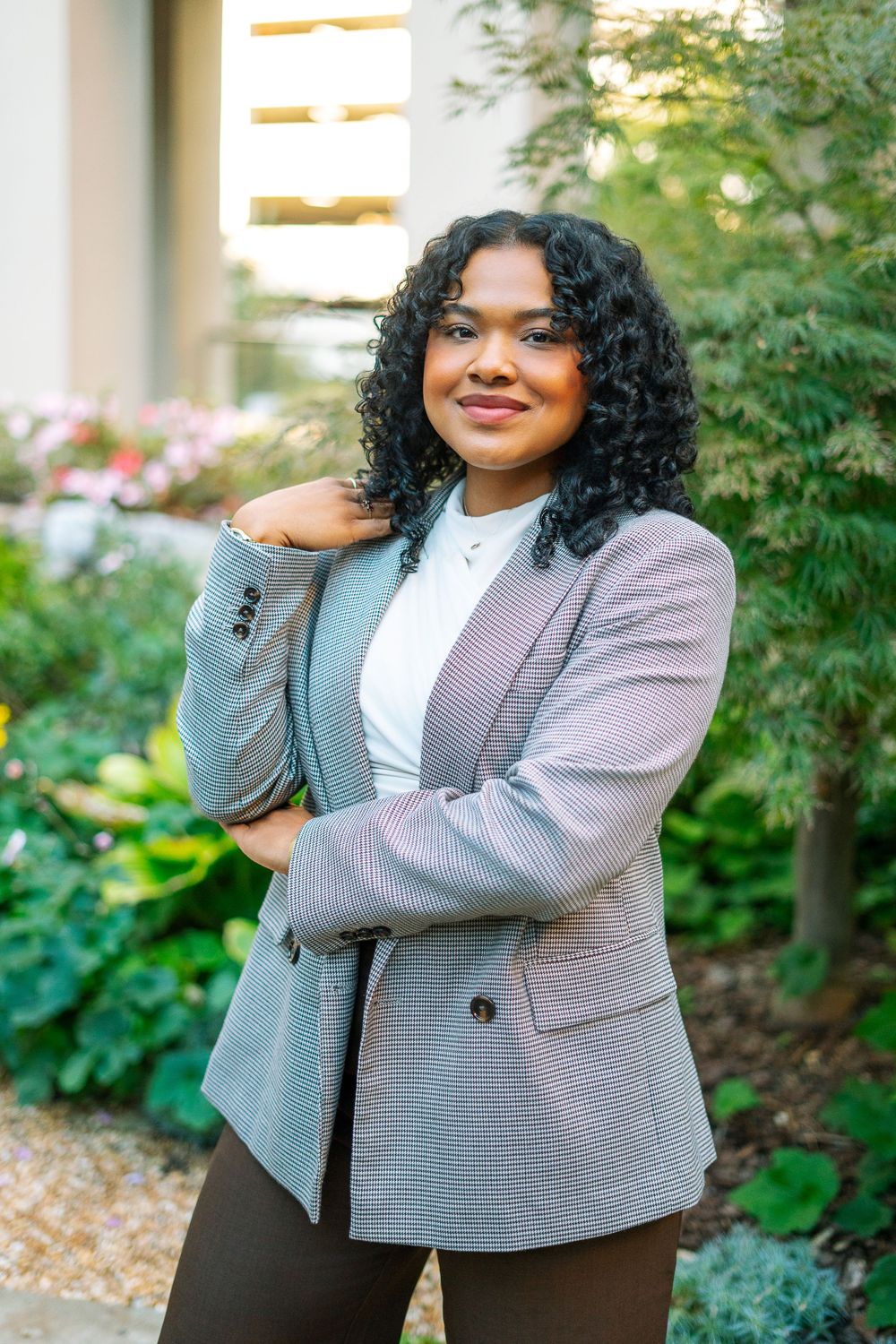 Young Black woman in plaid blazer posing confidently outdoors with greenery background.