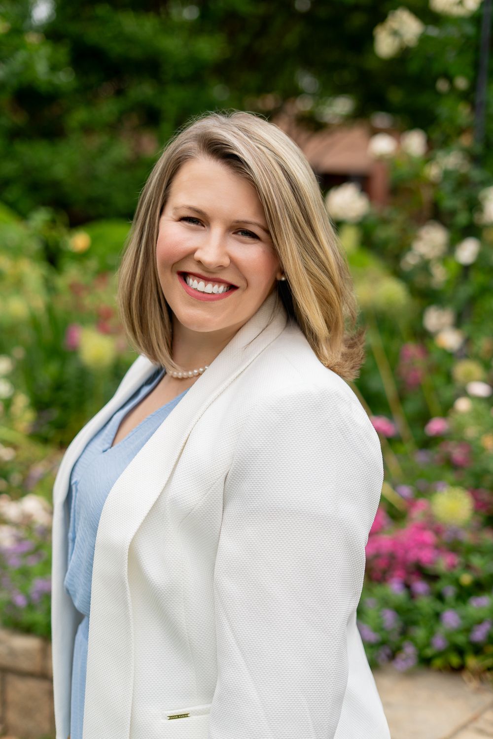 Professional blonde woman in white blazer smiling outdoors in a lush flower garden.