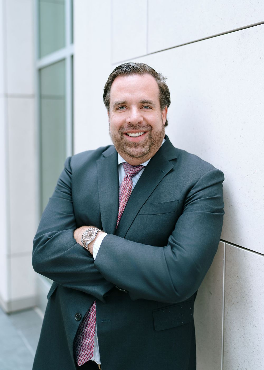 Smiling businessman in grey suit with pink tie leaning against wall with arms crossed.