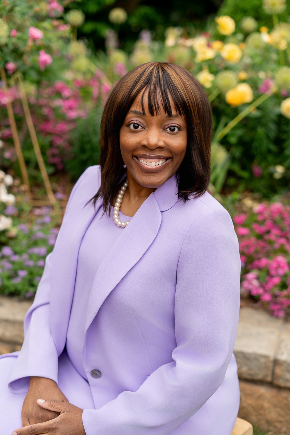 Elegant woman in lavender suit with pearl necklace smiling in vibrant flower garden.