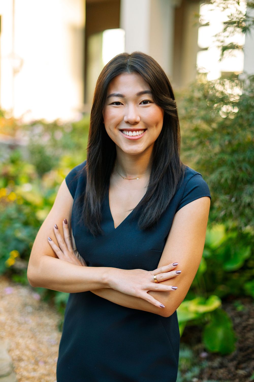 Smiling young Asian woman in navy dress with arms crossed outdoors in garden setting.
