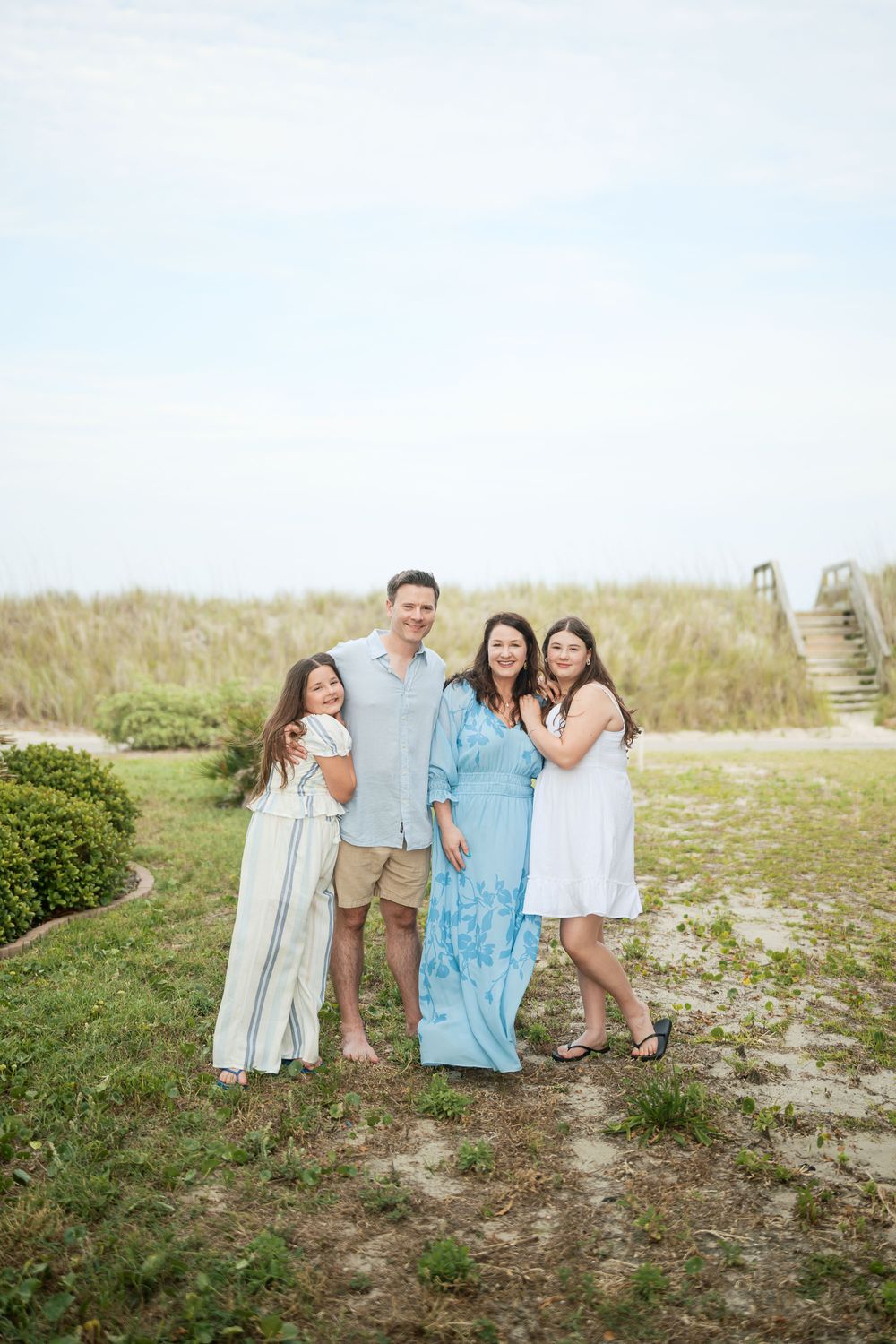Family of four smiling together on a grassy beach path, wearing light summer outfits near sandy dunes.