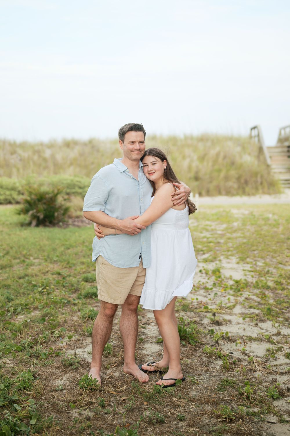 A man and young woman embrace while posing together on a sandy beach path, wearing casual summer attire.