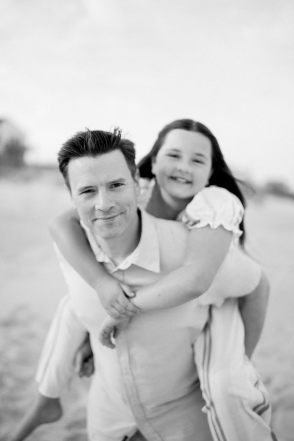 Black and white photo of a man giving a smiling girl a piggyback ride on a sandy beach.
