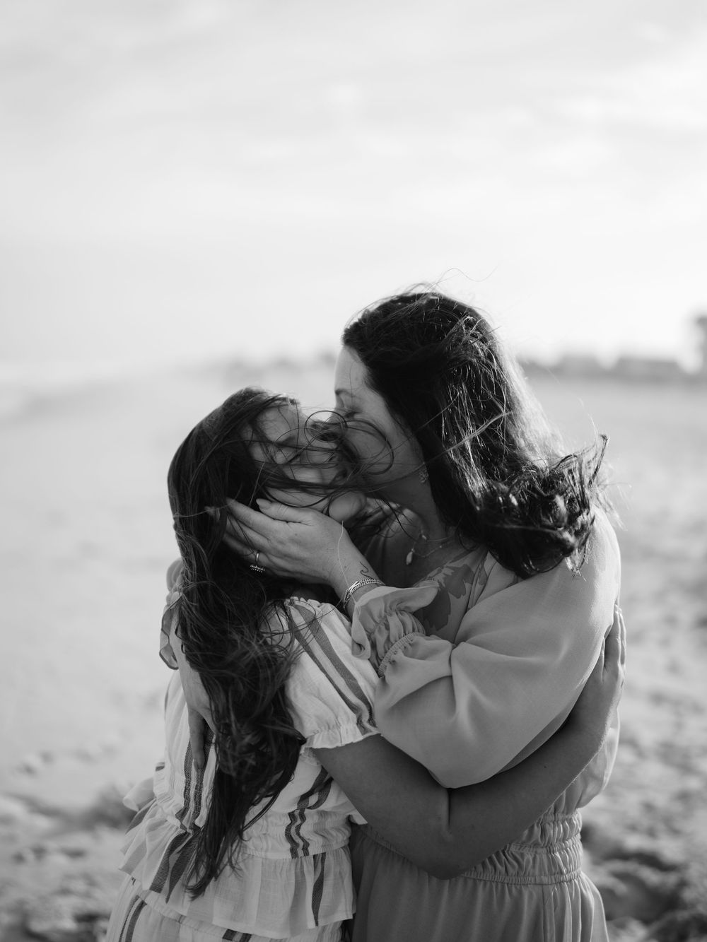 Black and white photo of a mother kissing her young daughter on a beach, both with long dark hair.