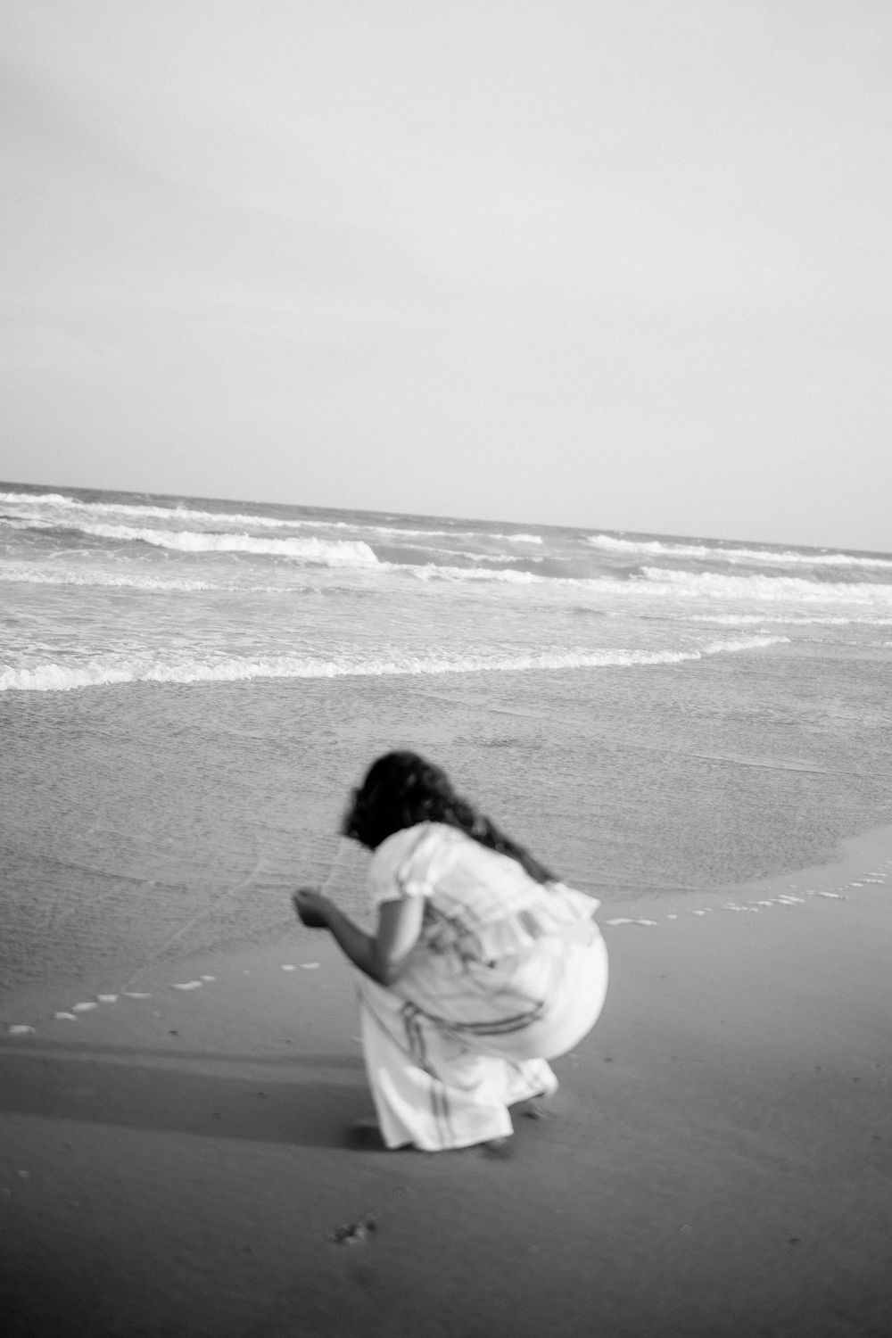 Black and white photo of a girl in a white dress crouching on a sandy beach with ocean waves in the background.