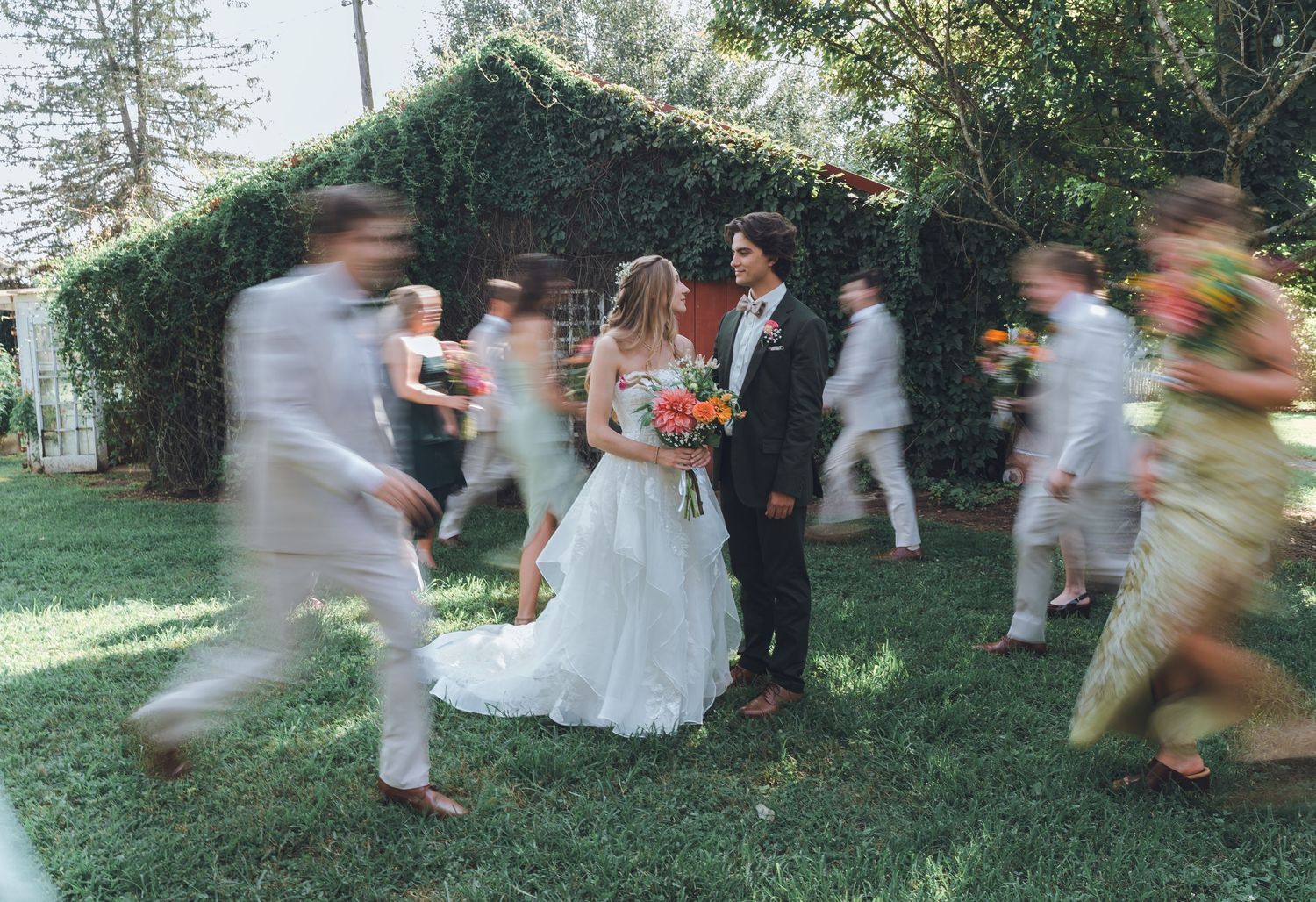 Bride and groom stand still amid blurred wedding party on lush green lawn beside ivy-covered brick wall.