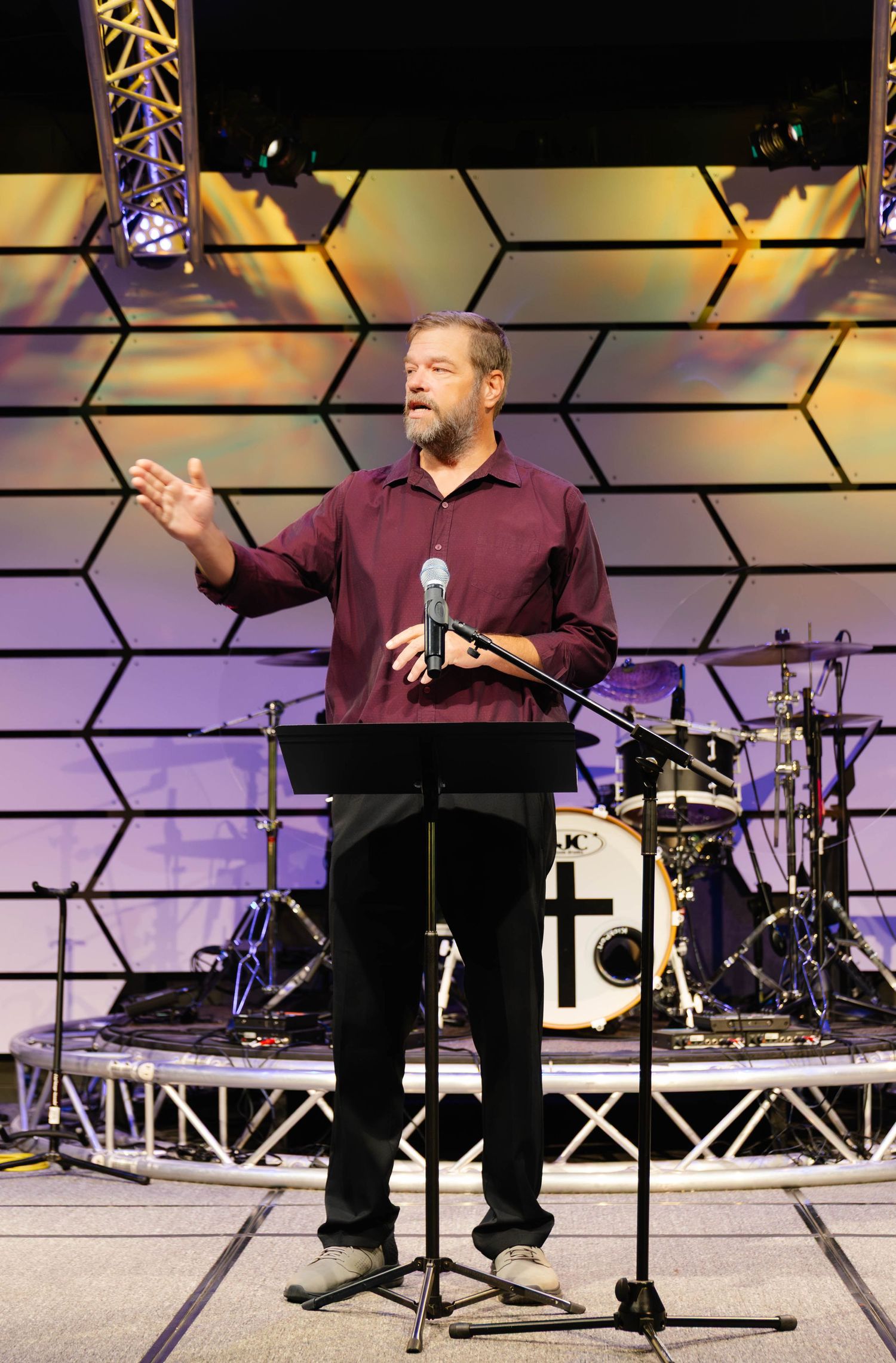 A bearded man in a maroon shirt speaks at a podium on a church stage with a cross, drum kit, and hexagonal backdrop.