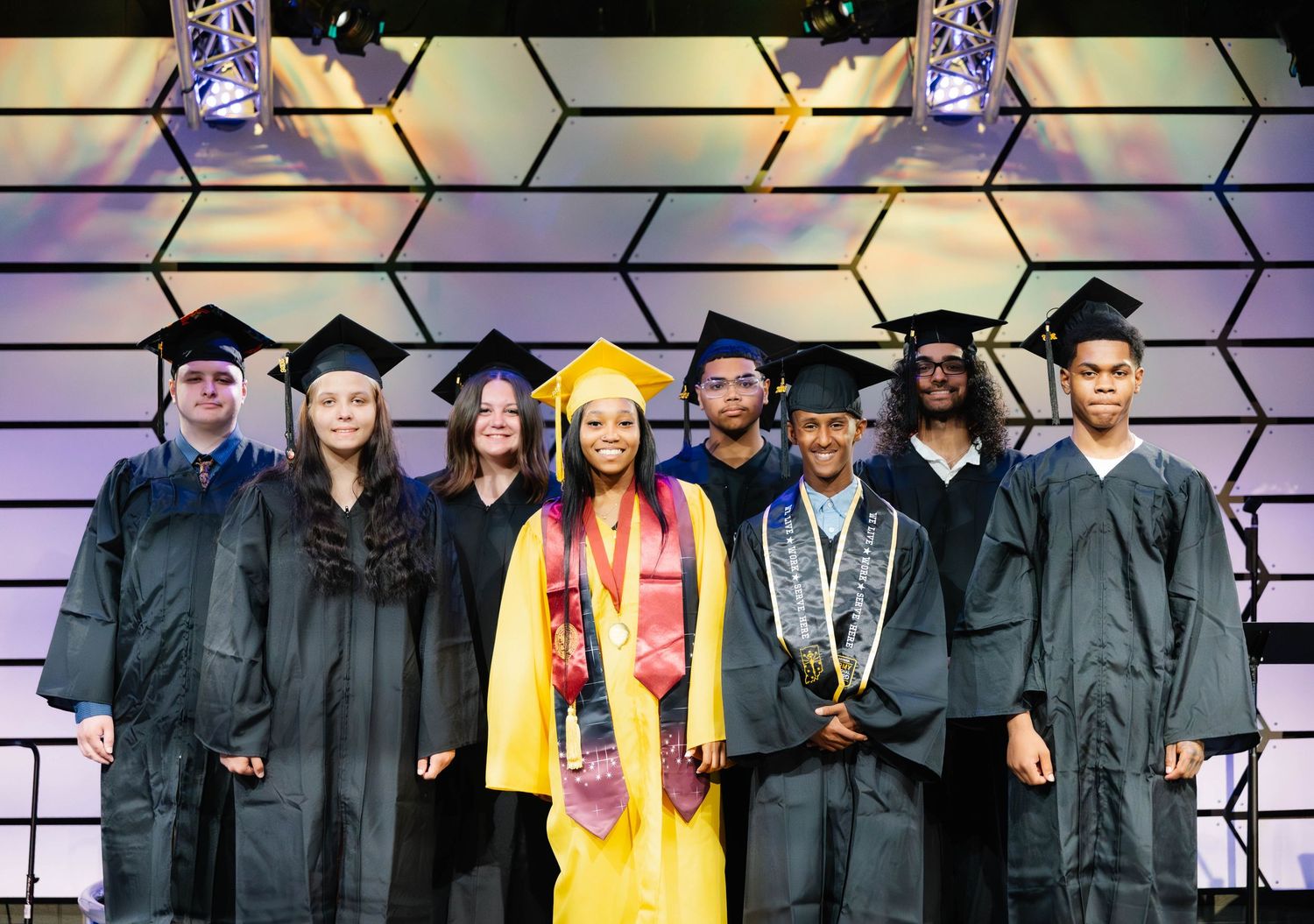 Group of graduates in caps and gowns posing on stage at graduation ceremony, one student in yellow regalia.