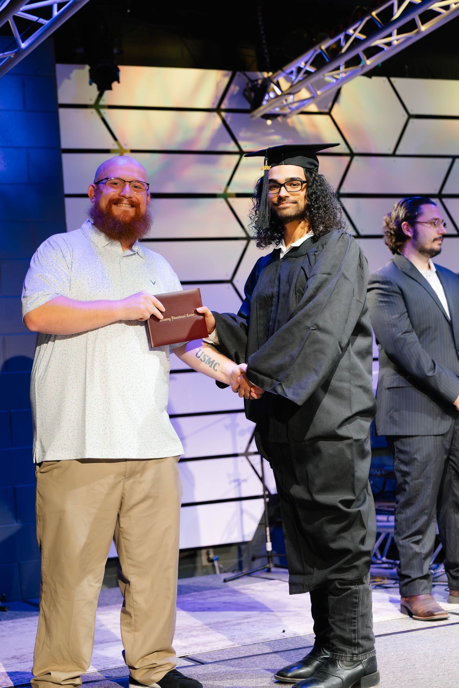 Graduate in cap and gown receives diploma on stage at graduation ceremony with hexagonal backdrop.
