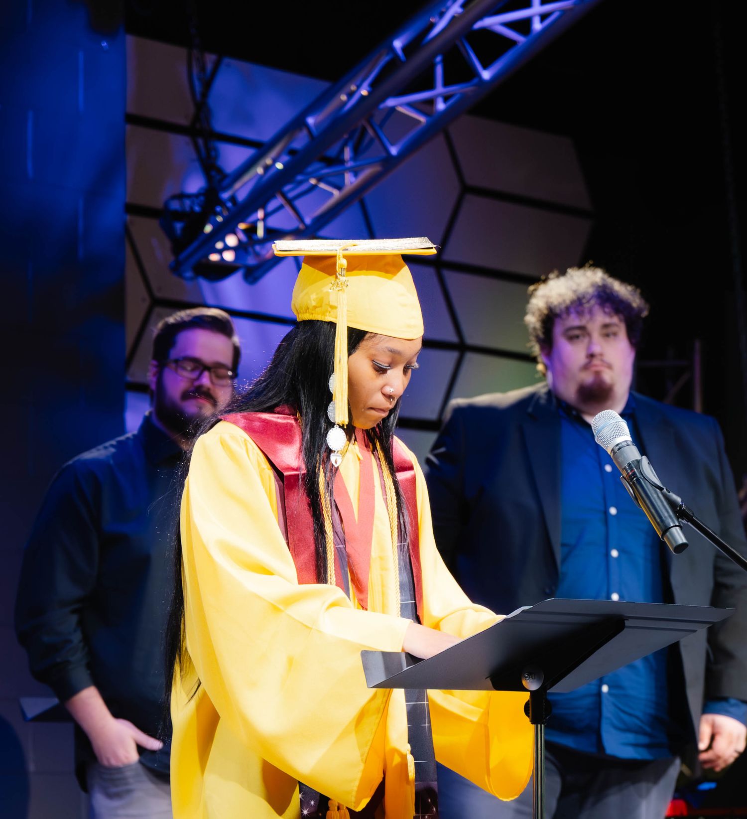 Graduate in yellow cap and gown reads at podium during graduation ceremony on stage.