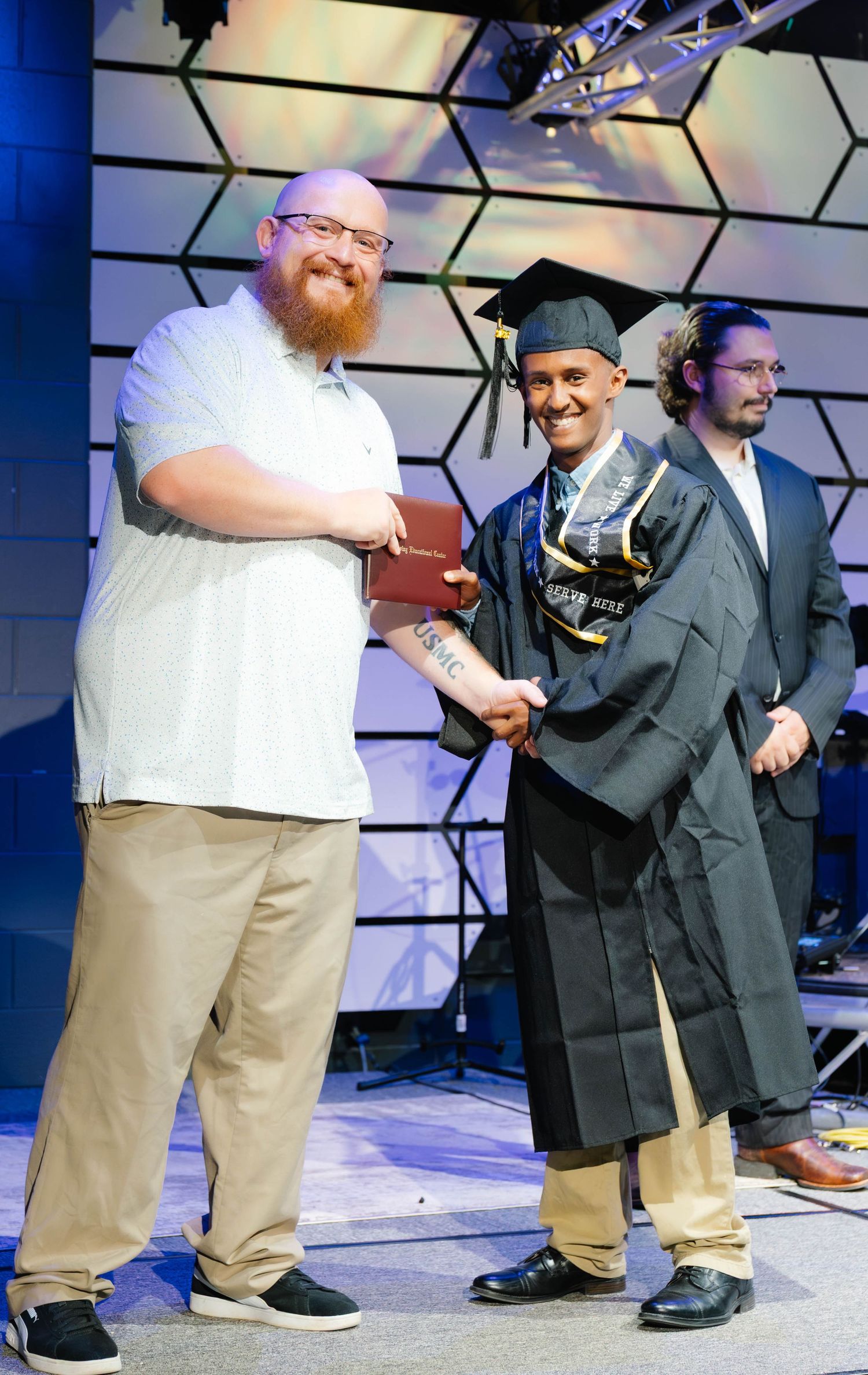 Graduate in cap and gown shaking hands and receiving diploma on stage at graduation ceremony.