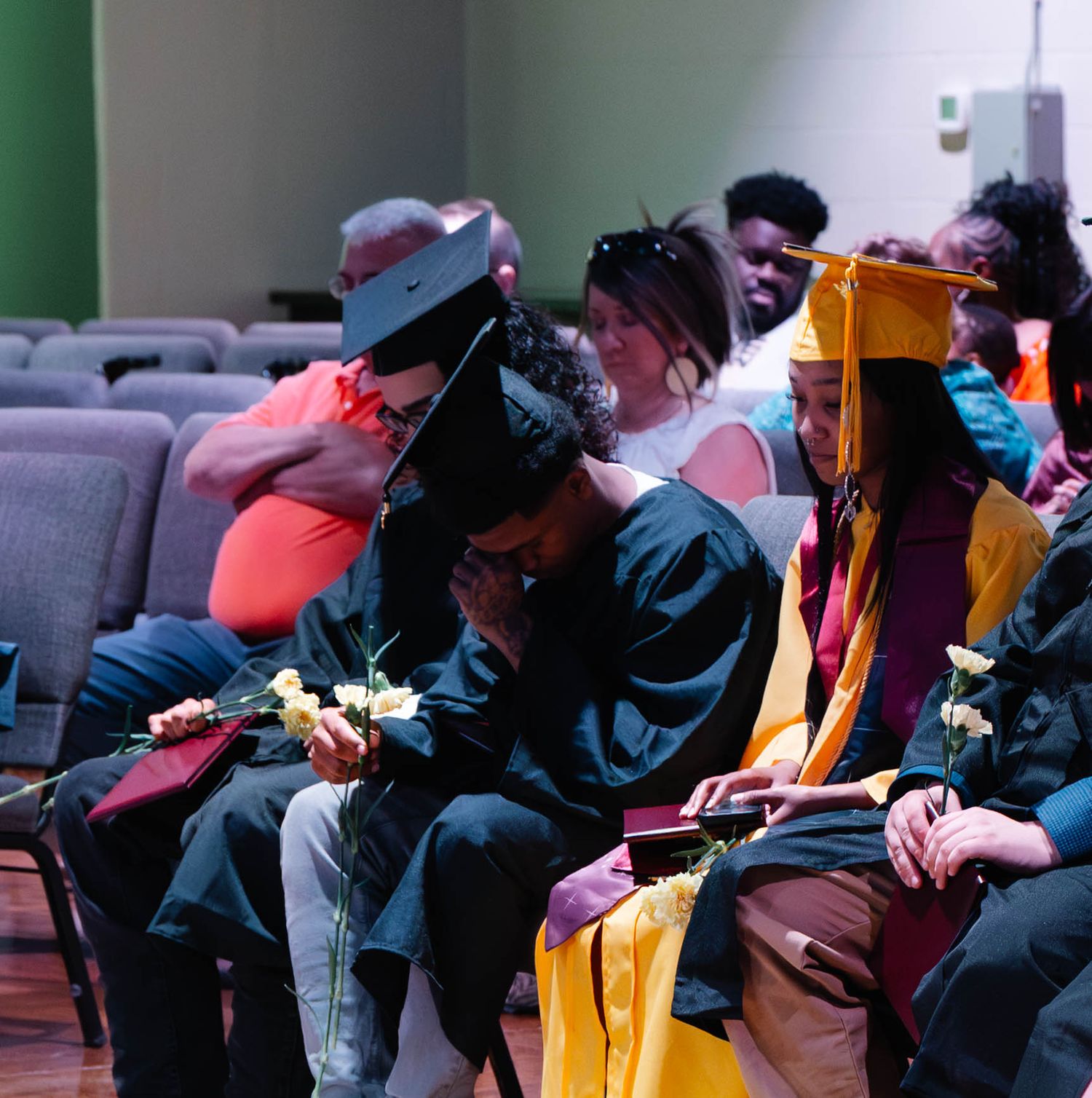 Graduates in caps and gowns share a moment at a graduation ceremony, holding flowers while seated in an auditorium.