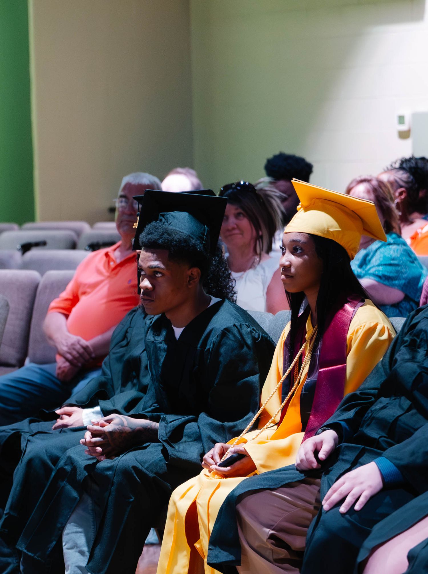 Two graduates in caps and gowns sit among guests at an indoor graduation ceremony.