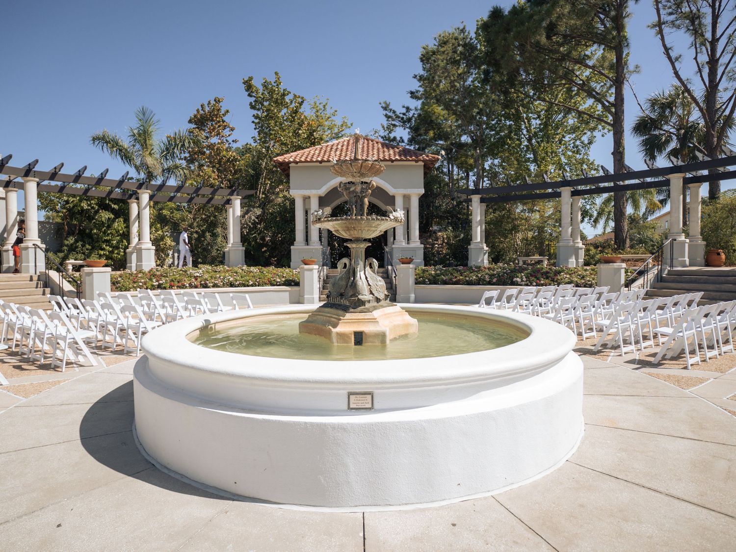 Elegant outdoor wedding venue with white chairs, fountain, pergola, and manicured gardens under a clear blue sky.