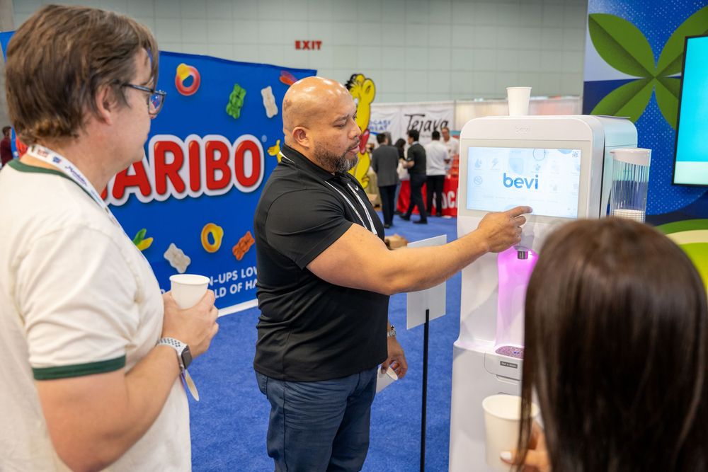 Man using a Bevi water dispenser kiosk at a trade show near a Haribo display booth.