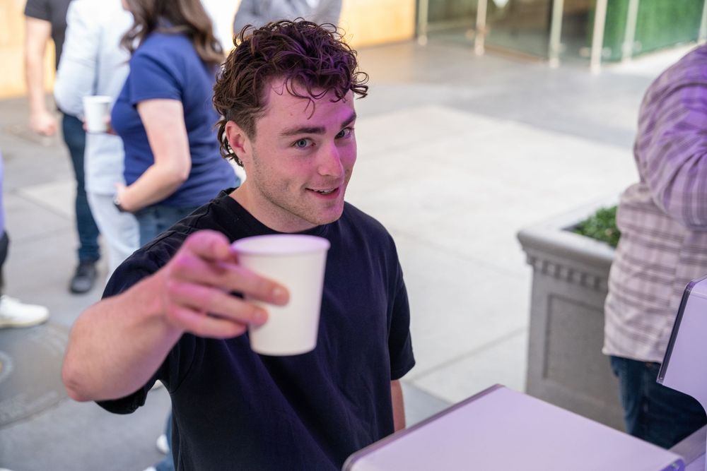 Young man in black t-shirt smiling and offering a white paper cup at an outdoor event.
