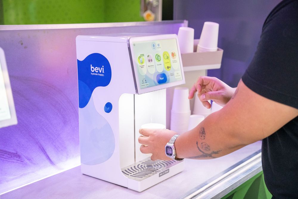 Person using a Bevi smart water dispenser machine with colorful touch screen interface in modern office.