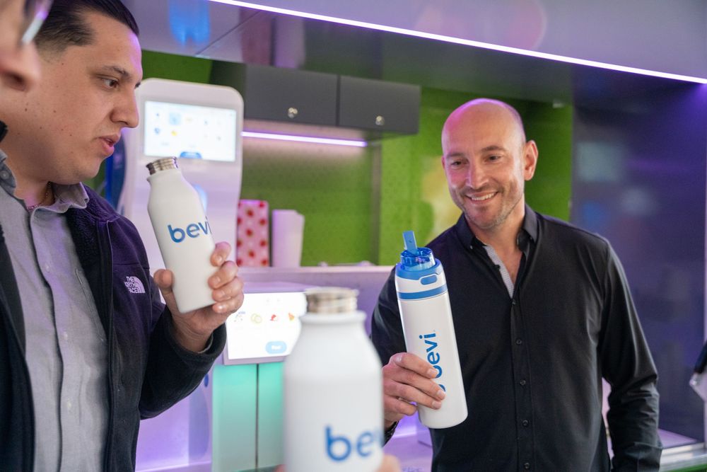 Two men smiling and holding Bevi branded reusable water bottles at a product demonstration event.