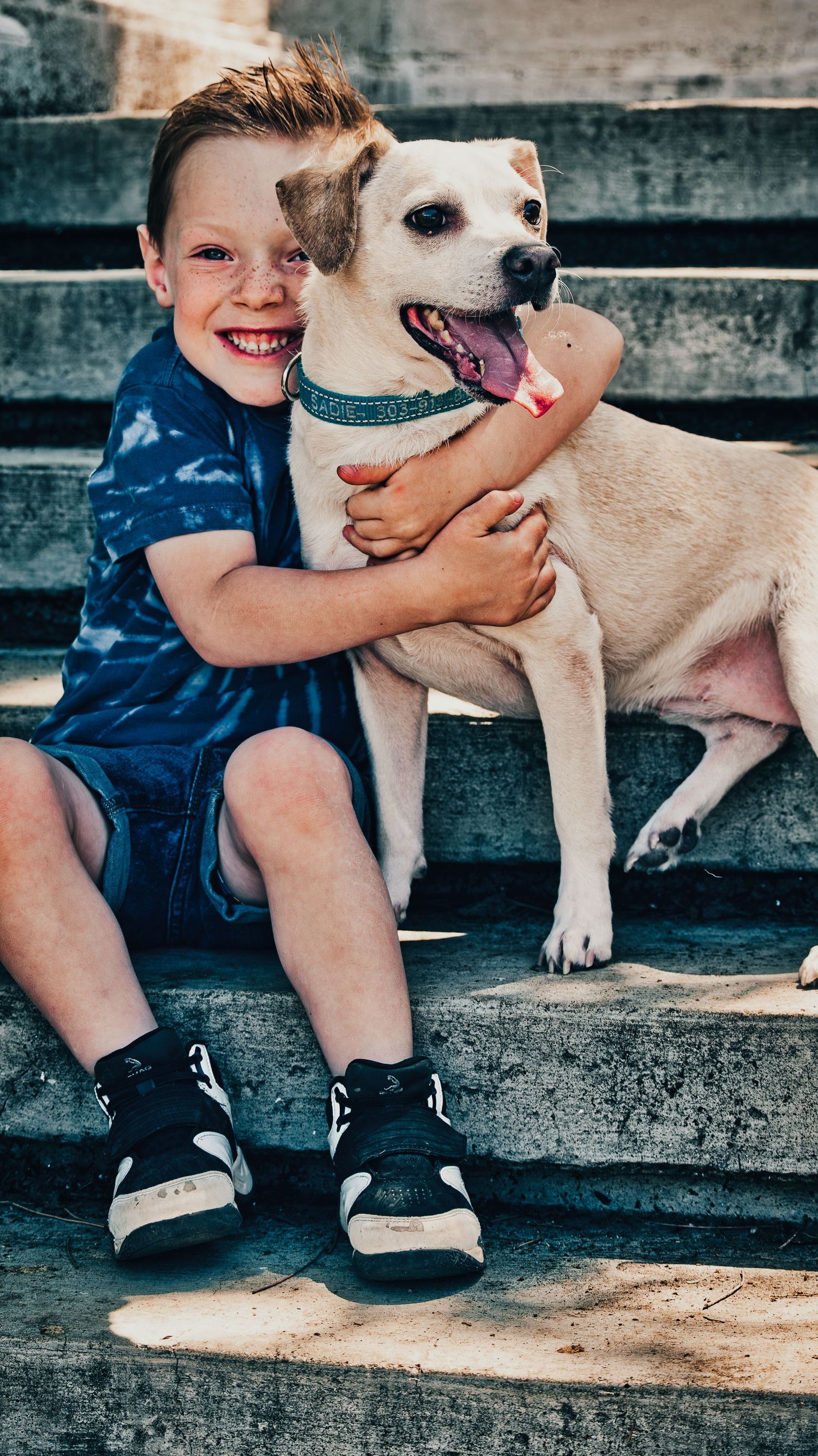 Smiling boy hugging a happy yellow Lab dog while sitting on concrete steps outdoors.
